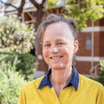 Student geologist Yeeda stand smiling on campus in front of a brick building and greenery. She wears her work uniform with her hair tied back.